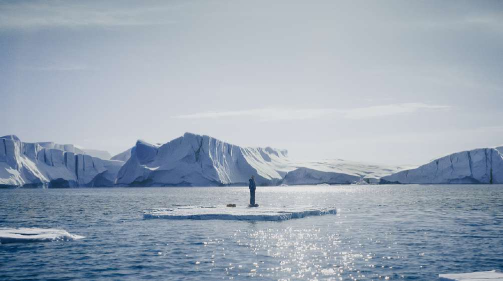 Emily standing alone on a small ice floe in an Arctic sea surrounded by distant glaciers.